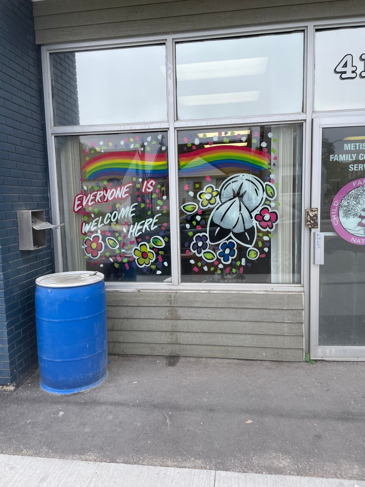 A blue barrel is sitting in front of a store window with a rainbow painted on it.