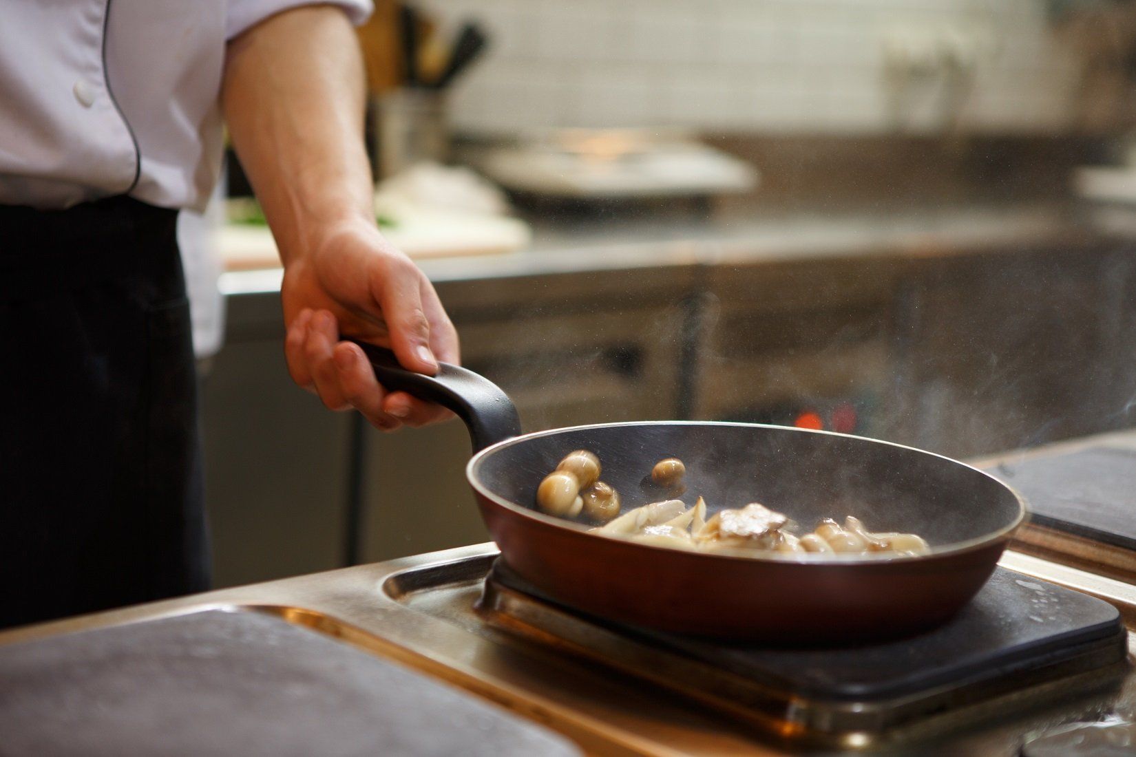 Mano dello chef che regge una padella di funghi che cuociono sul fornello di una cucina.