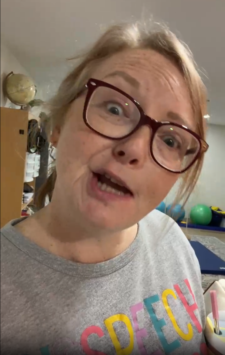 A person with blonde hair and glasses wearing a grey shirt with colorful text, looking into the camera in a room.