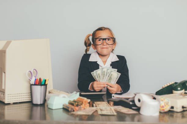 A person in a suit and glasses sits at a desk with an old computer and telephone, holding a fan of dollar bills.