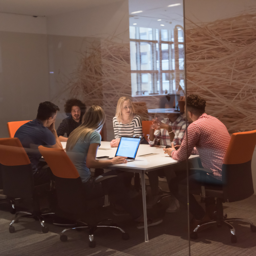 People at a table in an office, some looking at a laptop. Orange chairs, white table, glass walls.