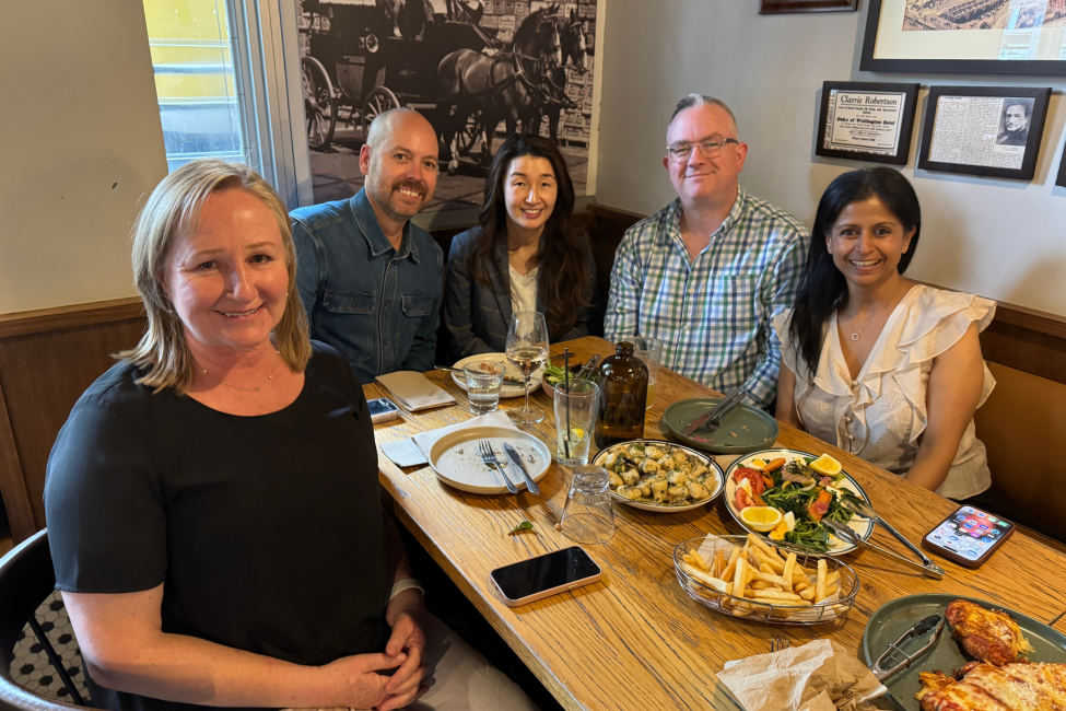 Five people seated around a restaurant table, smiling at the camera. Dishes of food and drinks are visible.