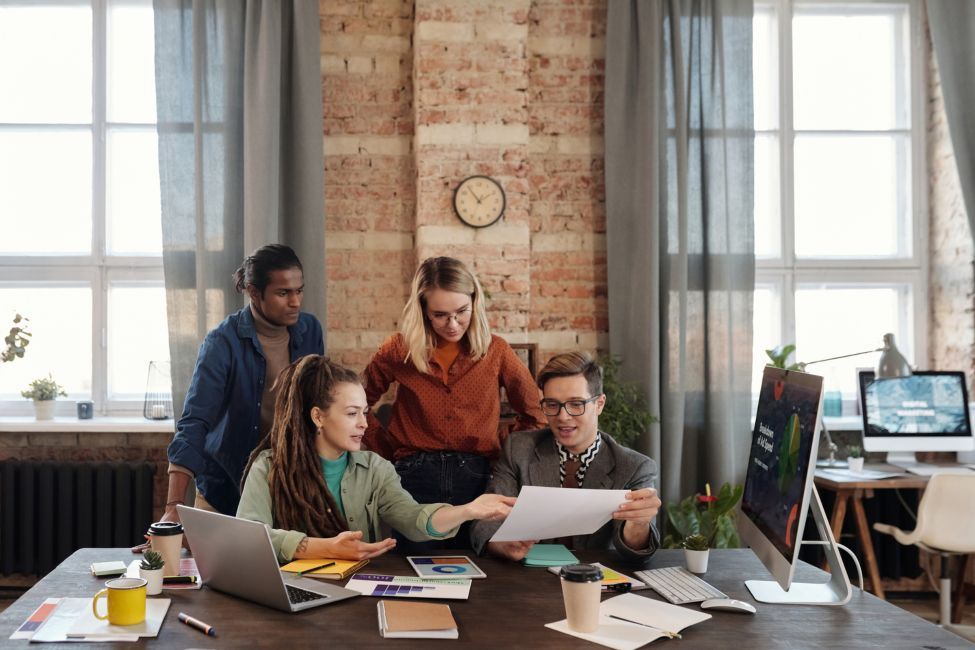 Four diverse coworkers collaborating around a desk with laptop, papers, and computer in a brick-walled office.
