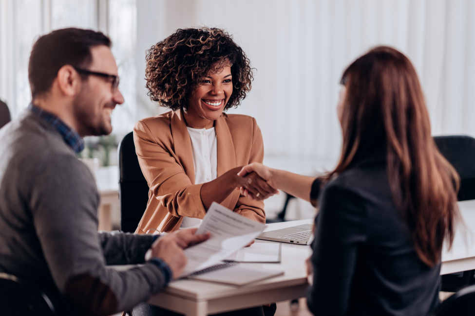Three coworkers in a meeting, smiling and shaking hands over documents at a desk