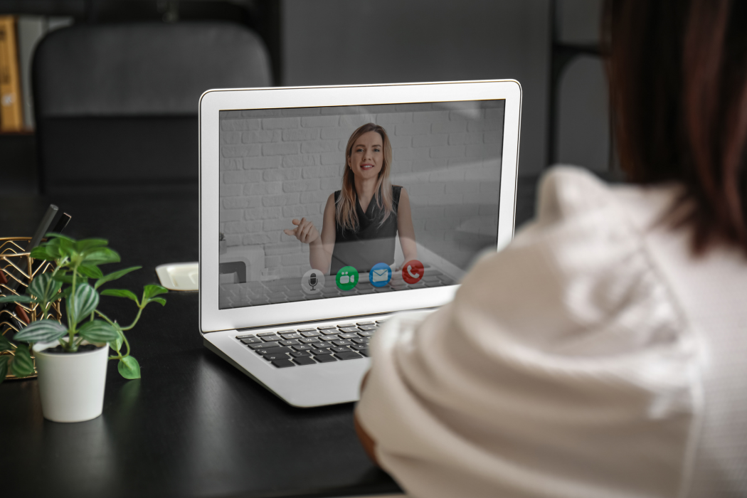 A person views a video call on a laptop screen, showing a presenter in a professional office setting.