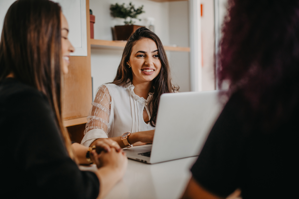 Three women at a table with a laptop, smiling. One in white shirt, two in dark tops, indoors.