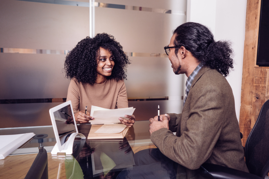 A person in a brown top smiles at a person in a brown blazer during a discussion. They are in an office.