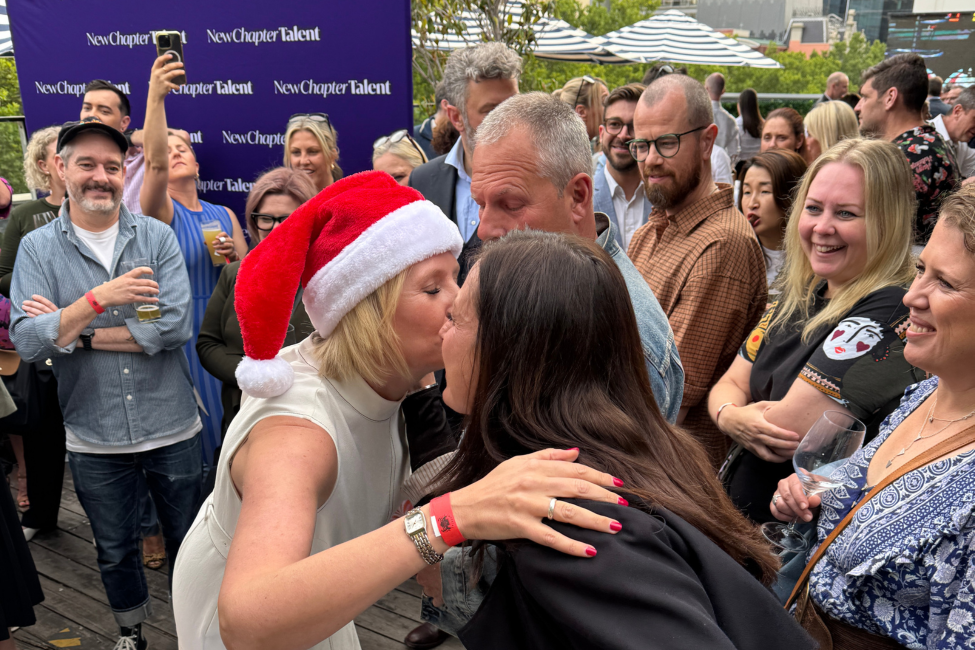 People at an outdoor gathering. Two women kissing, one wearing a Santa hat. Others smile.