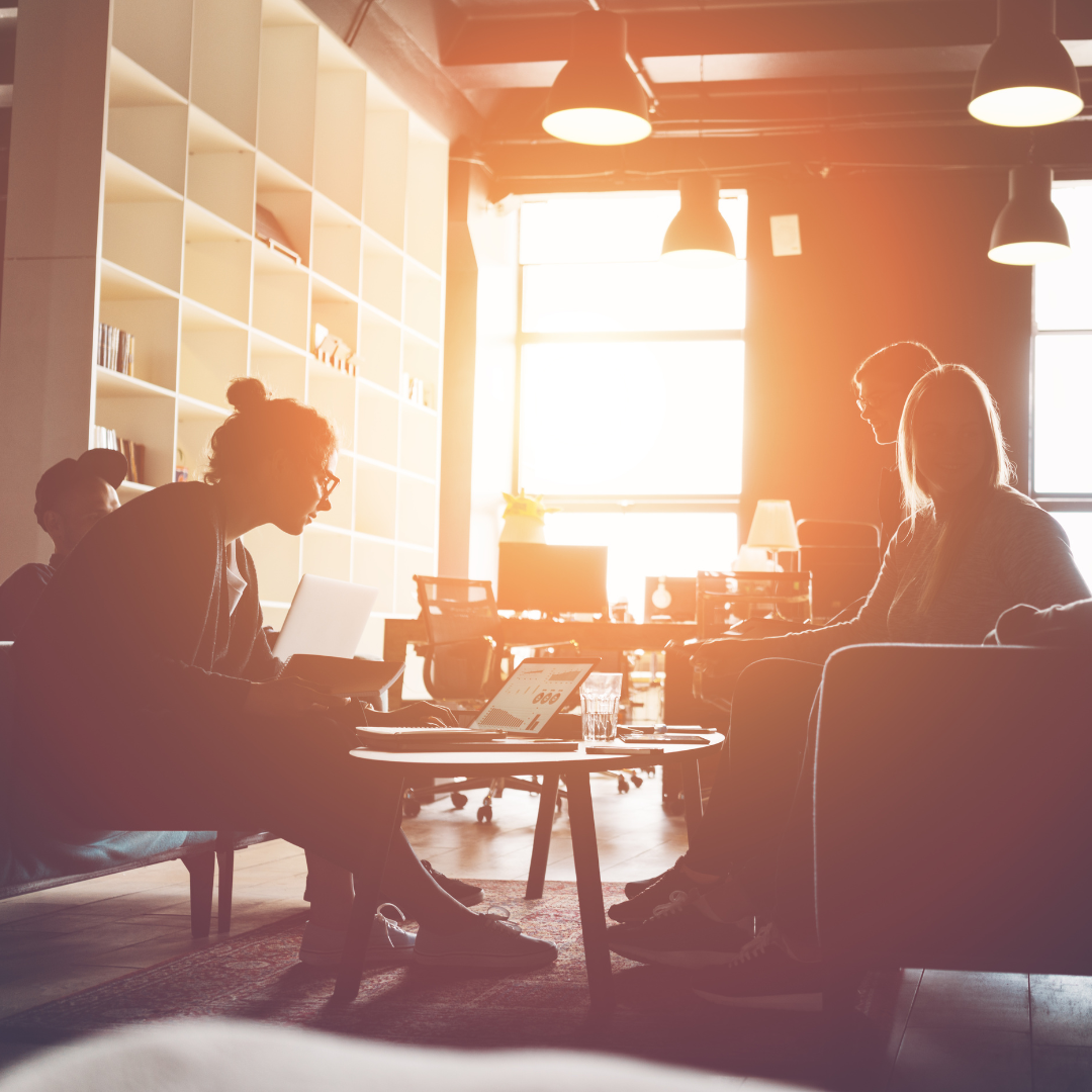 People in a well-lit office space, collaborating around a table with laptops. Sunlight streams in.