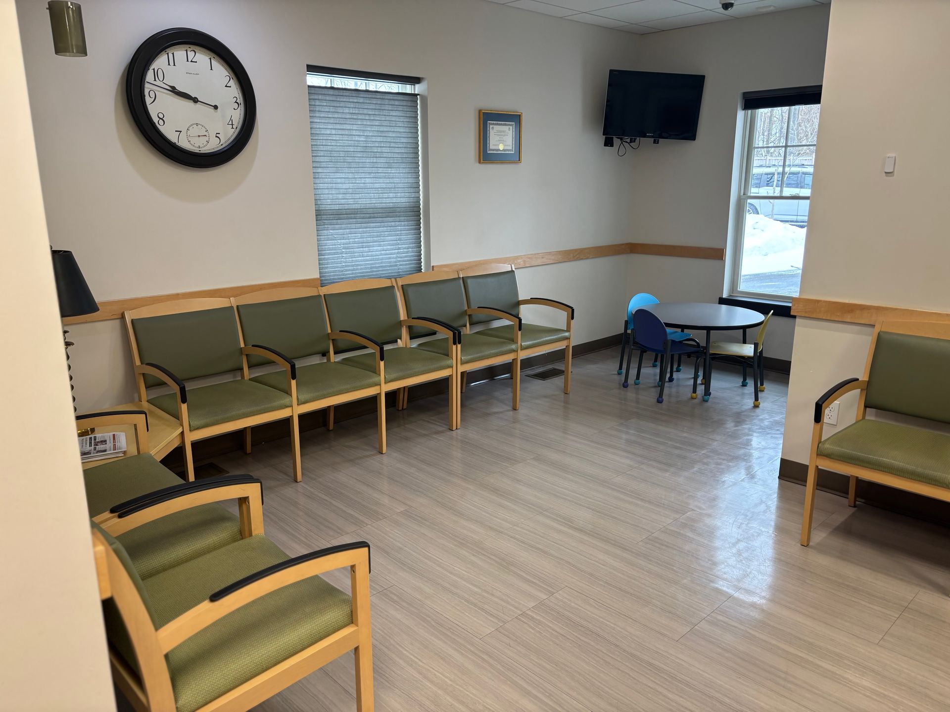 Waiting room with green chairs, clock, TV, small table, and window.