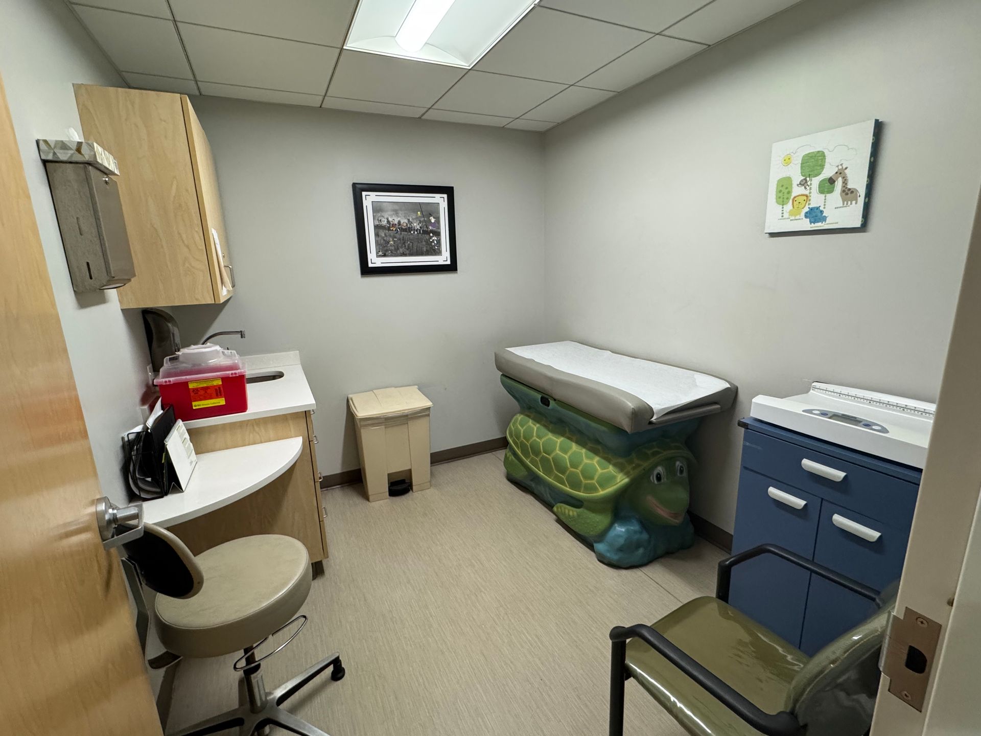 Examination room with light beige walls, examining table, storage, and small chair.