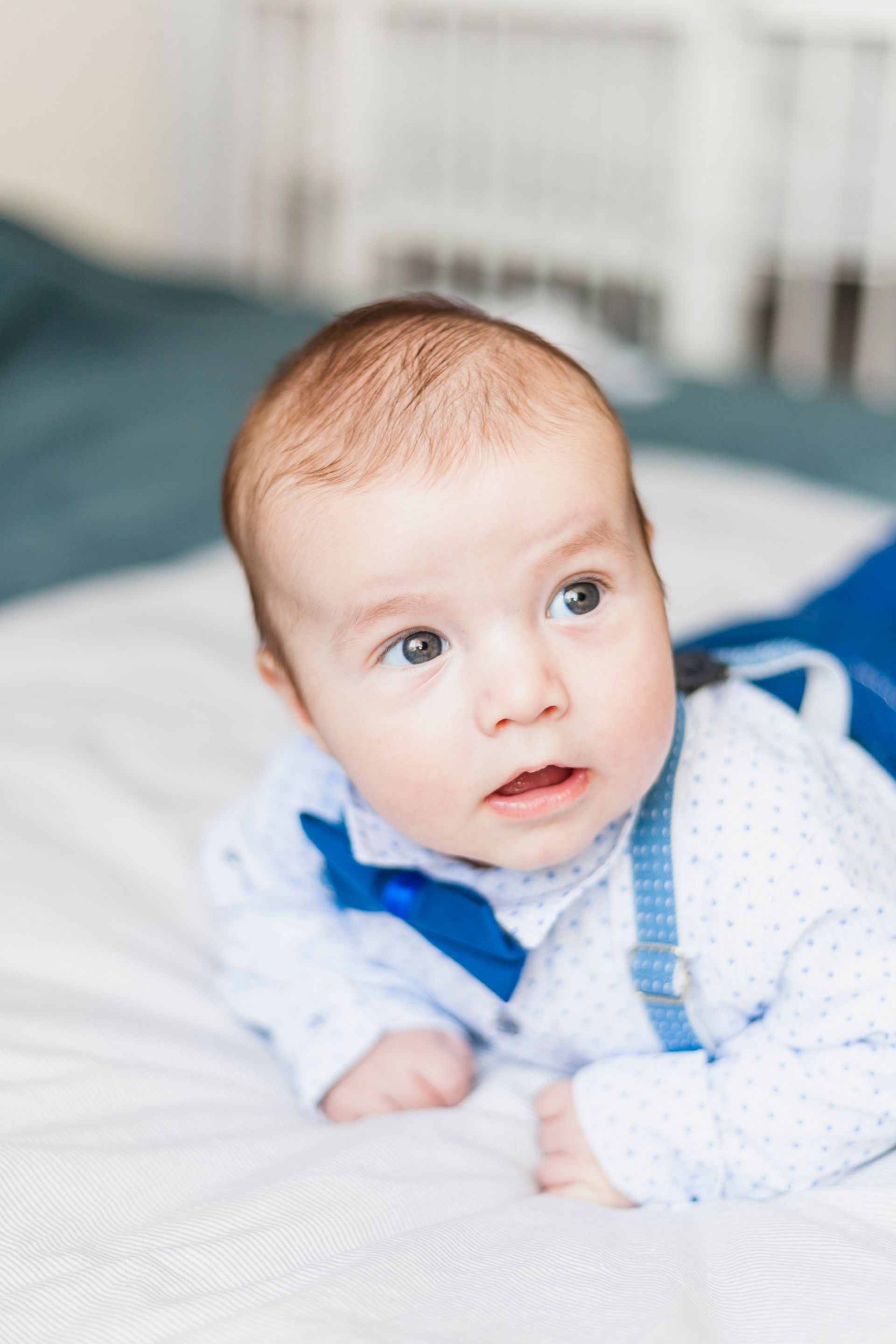 Baby in white shirt and blue suspenders, looking up on a white bed.