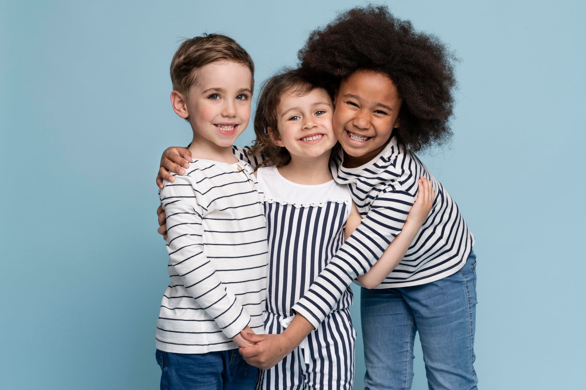 Three smiling children in striped shirts hug against a blue backdrop.