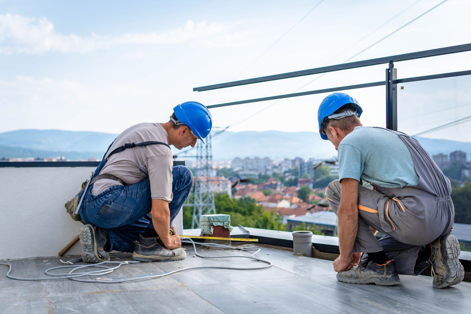Two men wearing hard hats and coveralls are on a roof, performing a flat roof repair. Two men wearing hard hats and coveralls are on a roof, performing a flat roof repair.