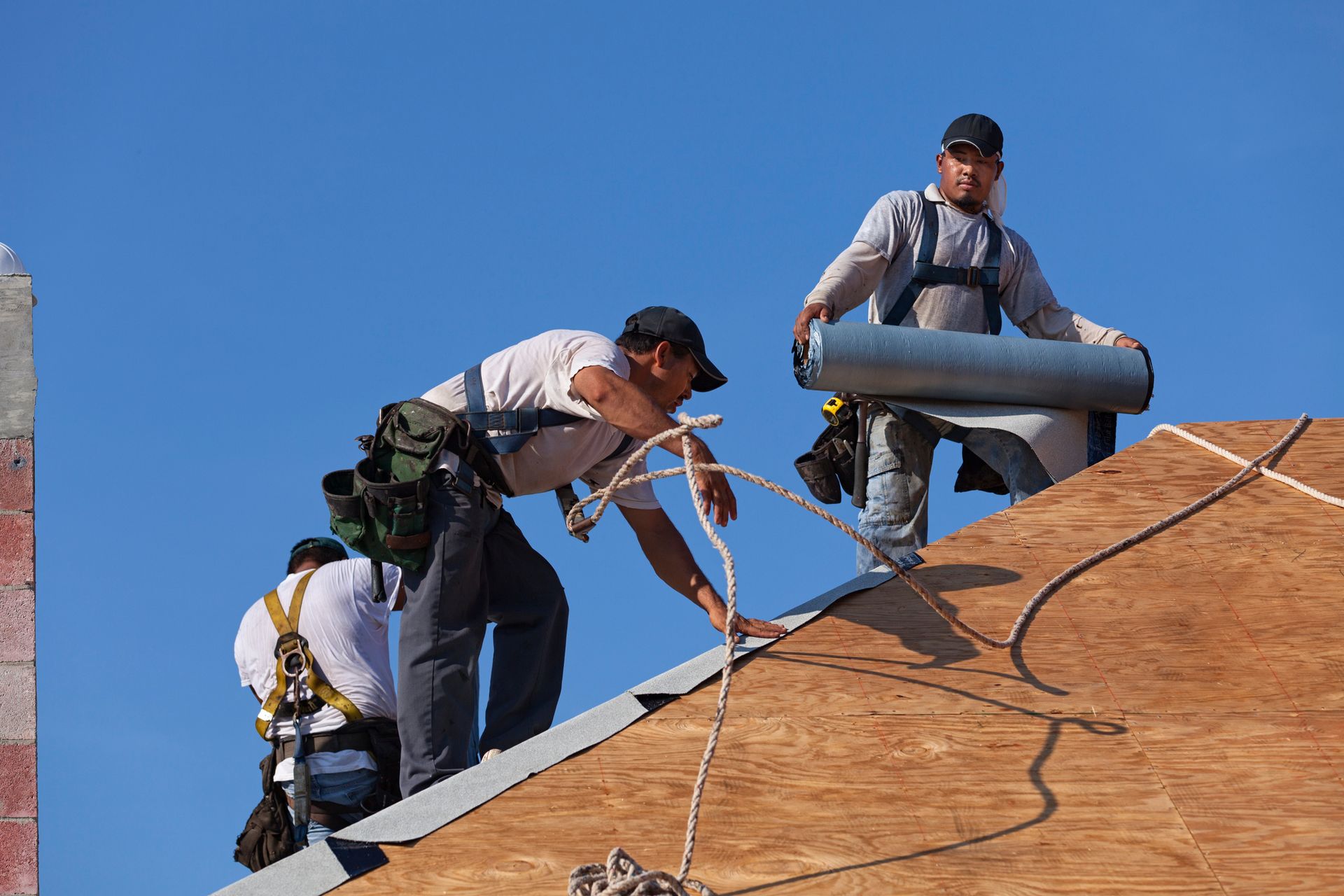 Three roofers installing roofing material on a wooden roof under a blue sky.