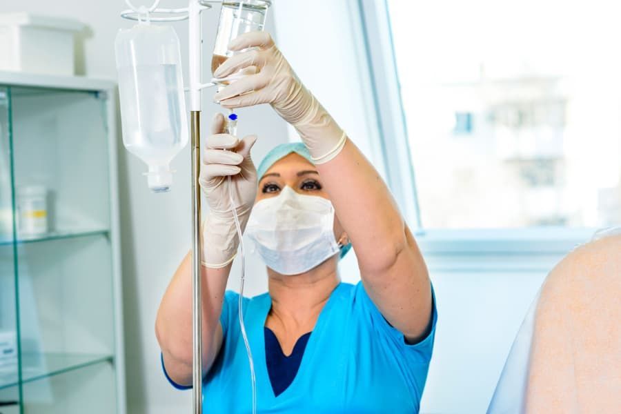 Nurse in scrubs and mask adjusting an IV drip in a medical setting.