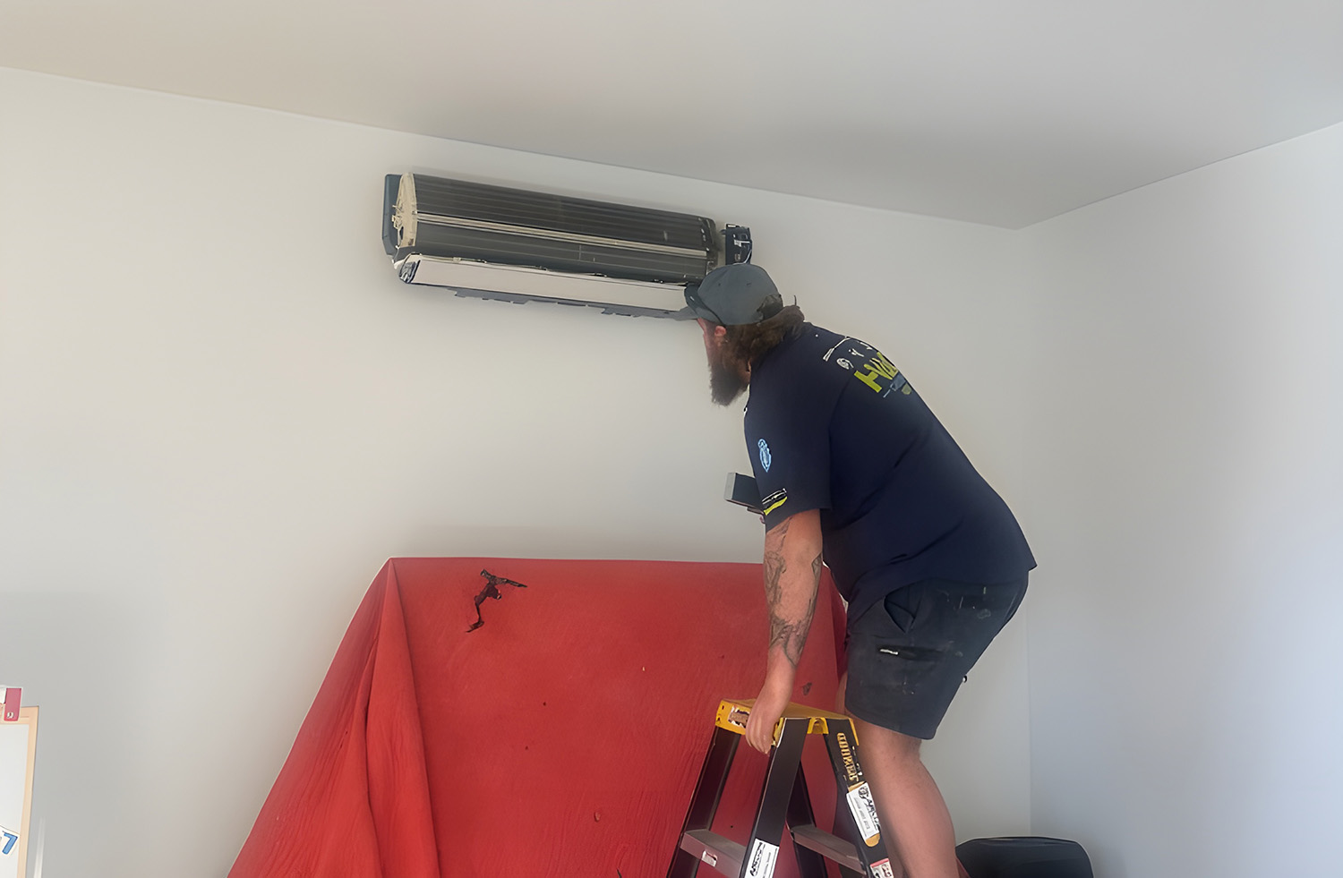 A Service Technician On A Stepladder Cleaning A Wall-Mounted Air Conditioning Unit