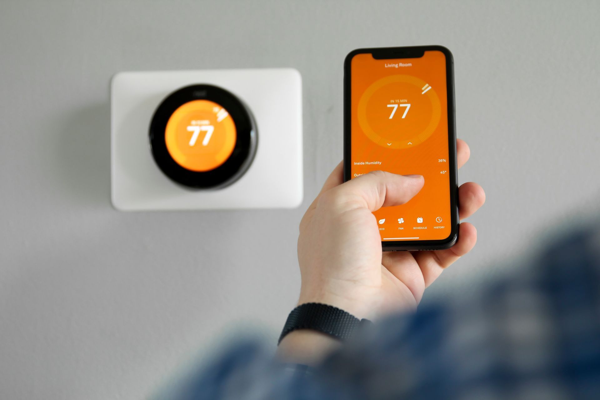 Person using smartphone to control a smart thermostat, both displaying 77 degrees.