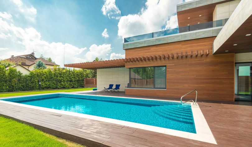 Modern house with a swimming pool on a sunny day, blue water, wooden deck, and green lawn.
