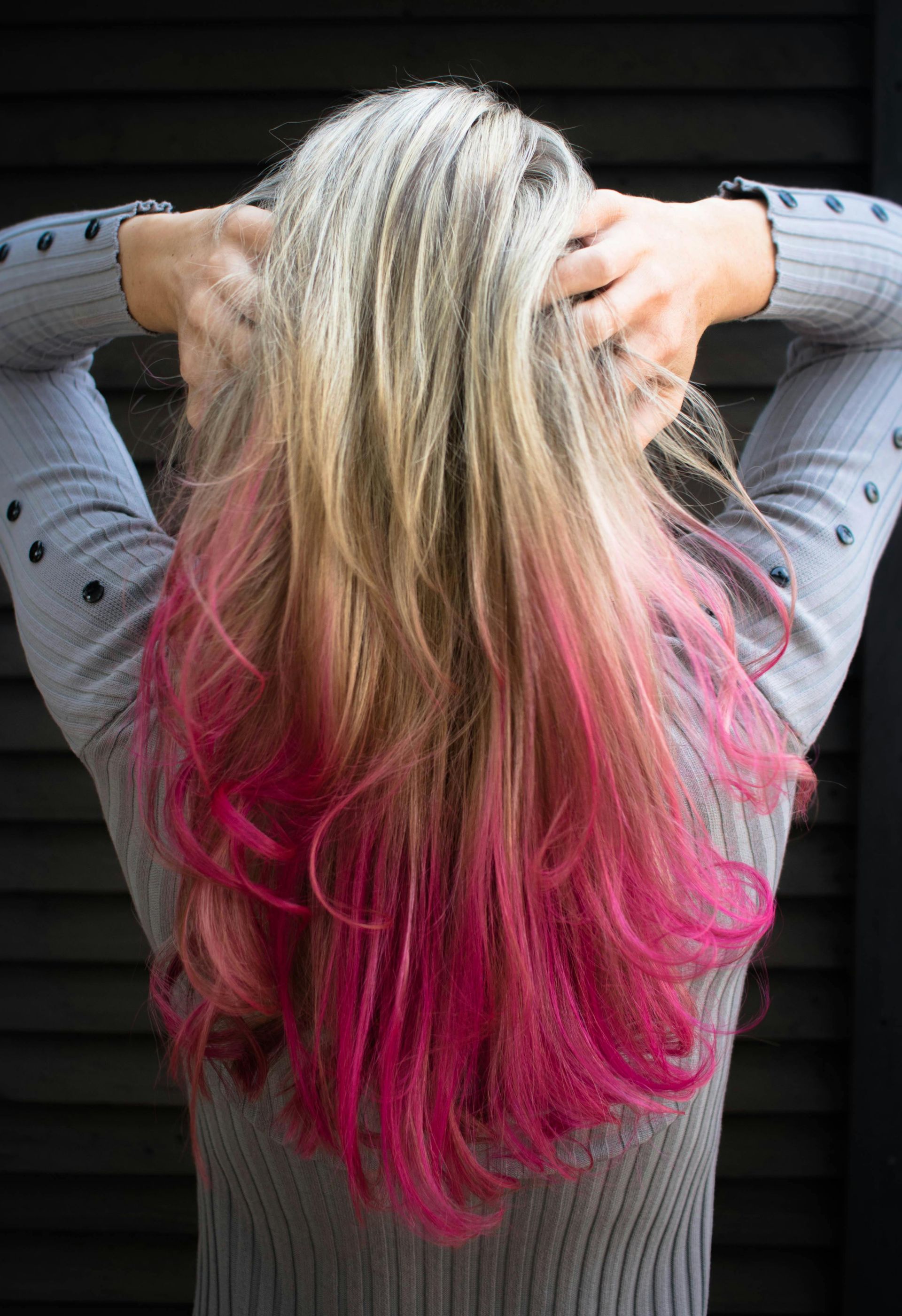 Person with long, wavy blonde hair with bright pink dip-dye, hands in hair, gray top, black background.