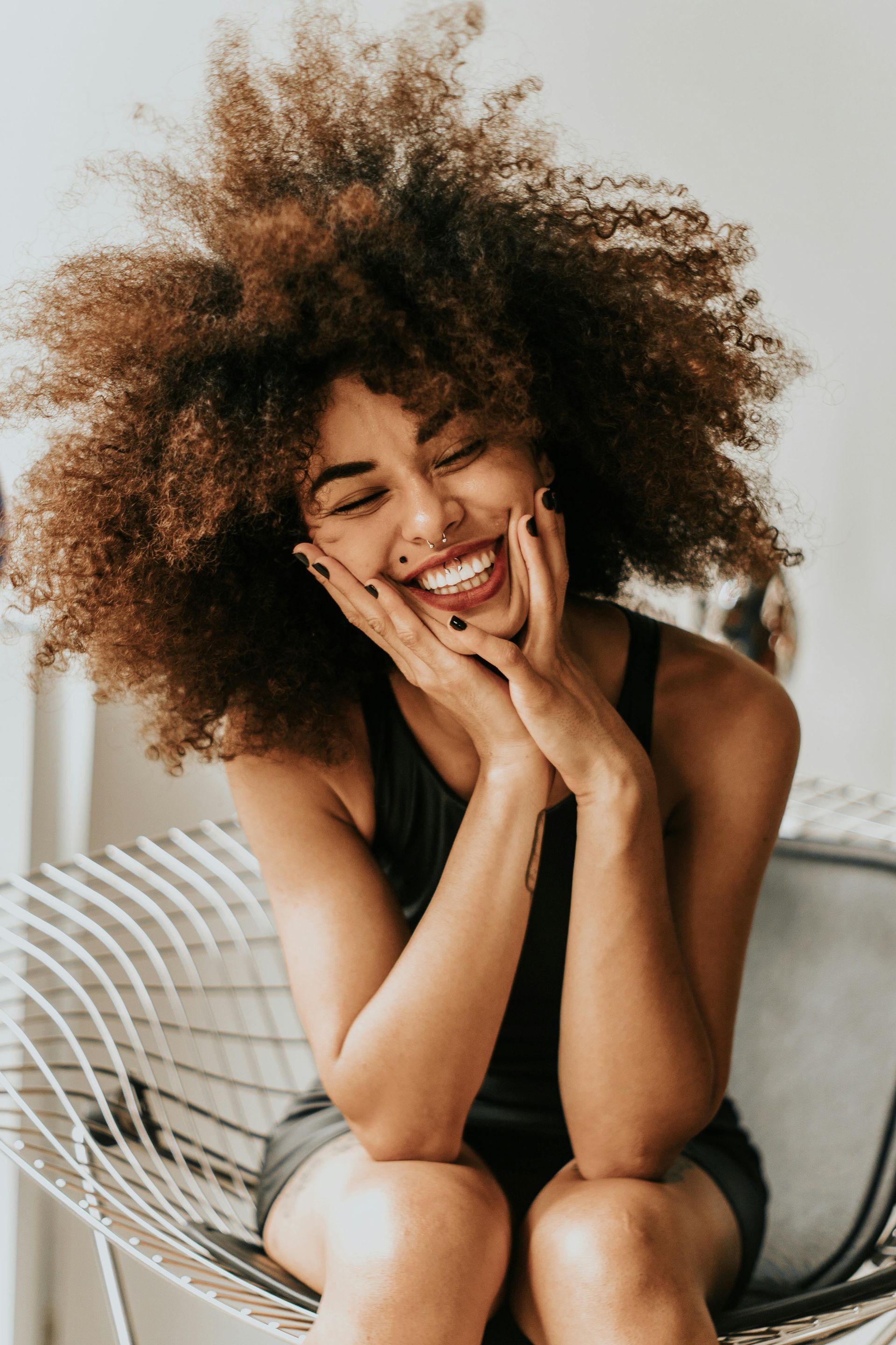 Woman with large curly hair smiles, holding cheeks, sitting in a metal chair.