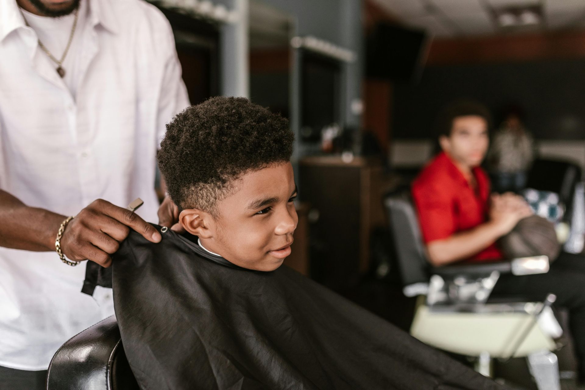 Person in barber chair, getting a haircut. Barber drapes cape. Another person in background.