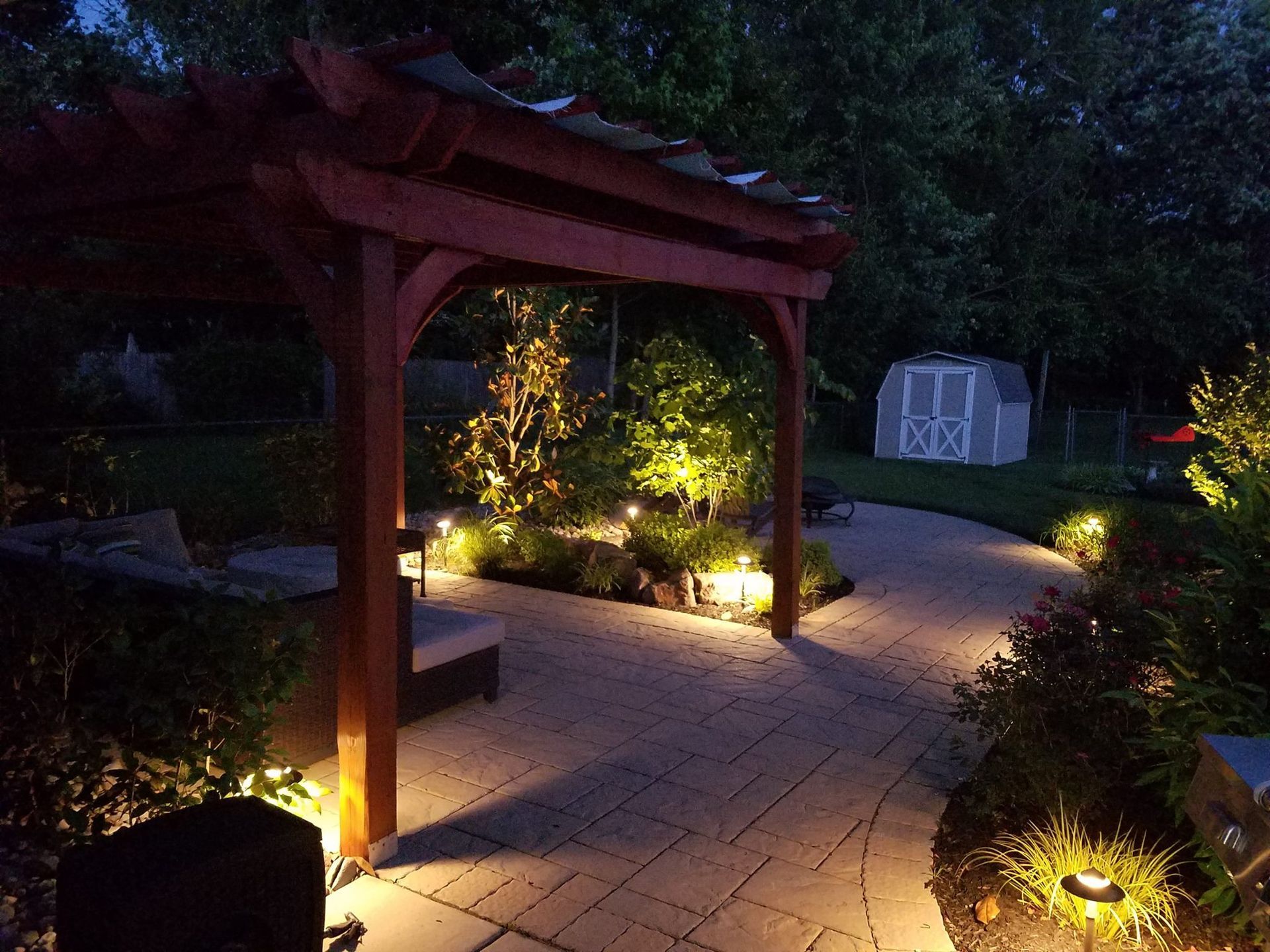 A pergola is lit up at night in a backyard with a shed in the background.