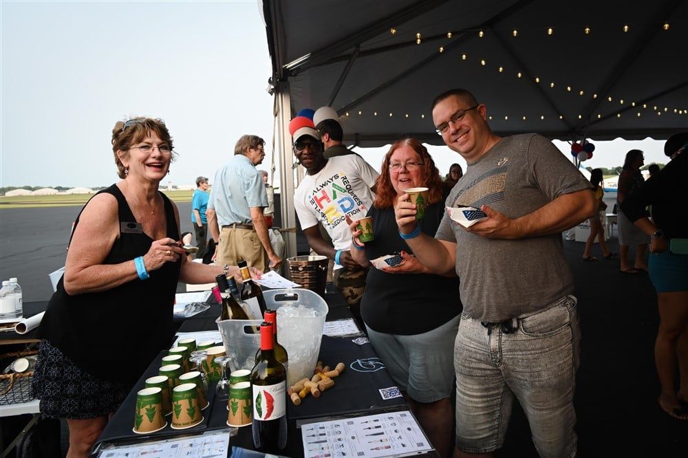 A group of people are standing around a table with bottles of wine.