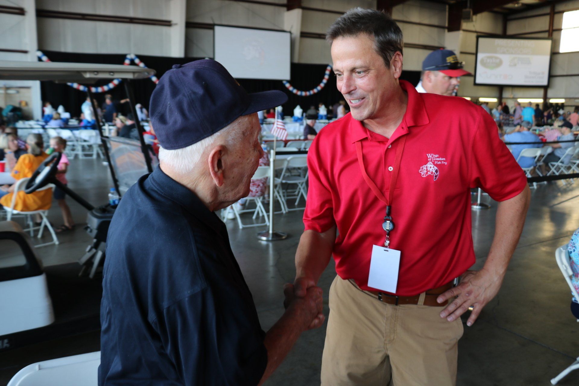 A man in a red shirt is shaking hands with an older man