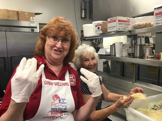 Two women wearing aprons and gloves are standing in a kitchen.