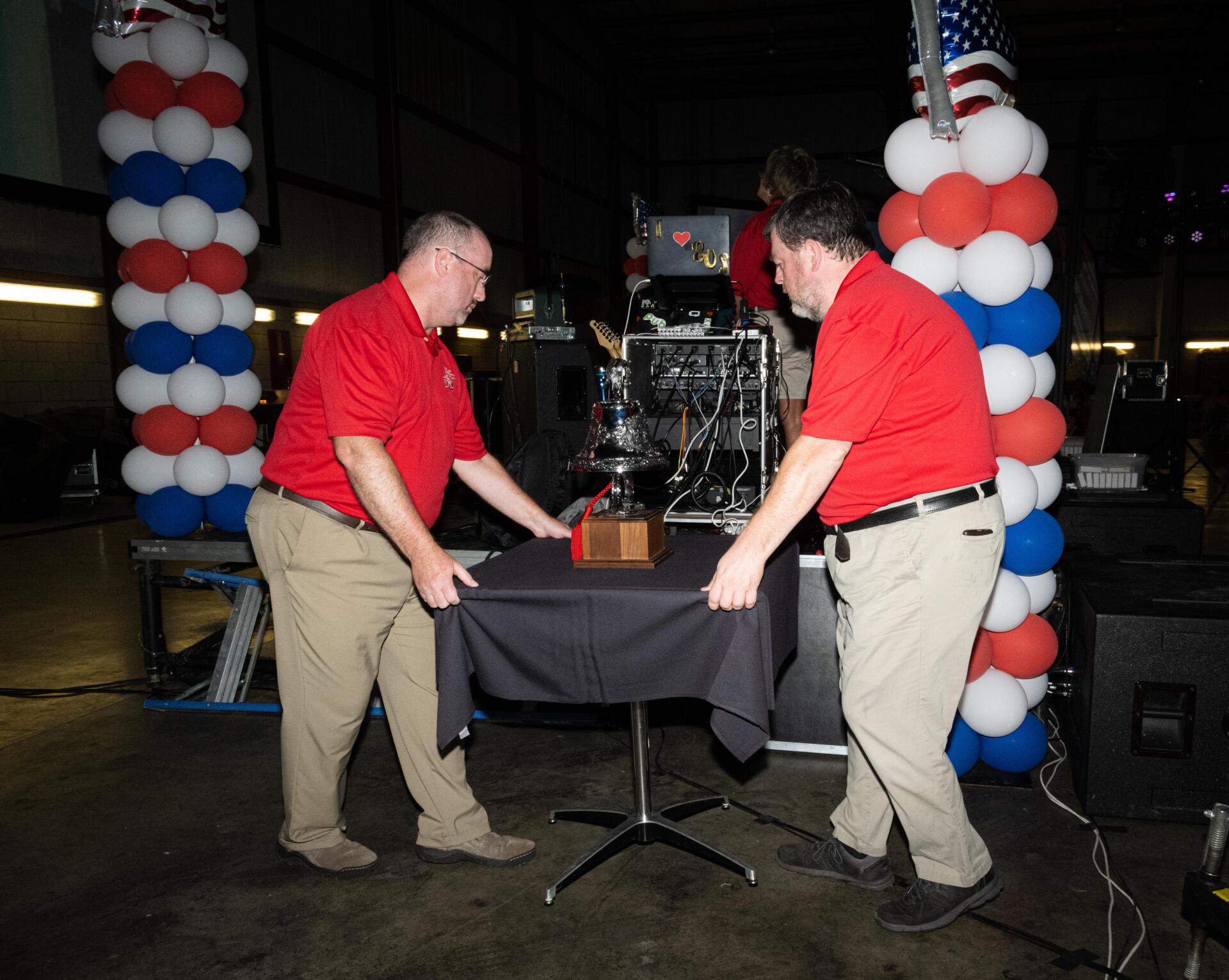 Two men in red shirts are standing around a table with balloons in the background