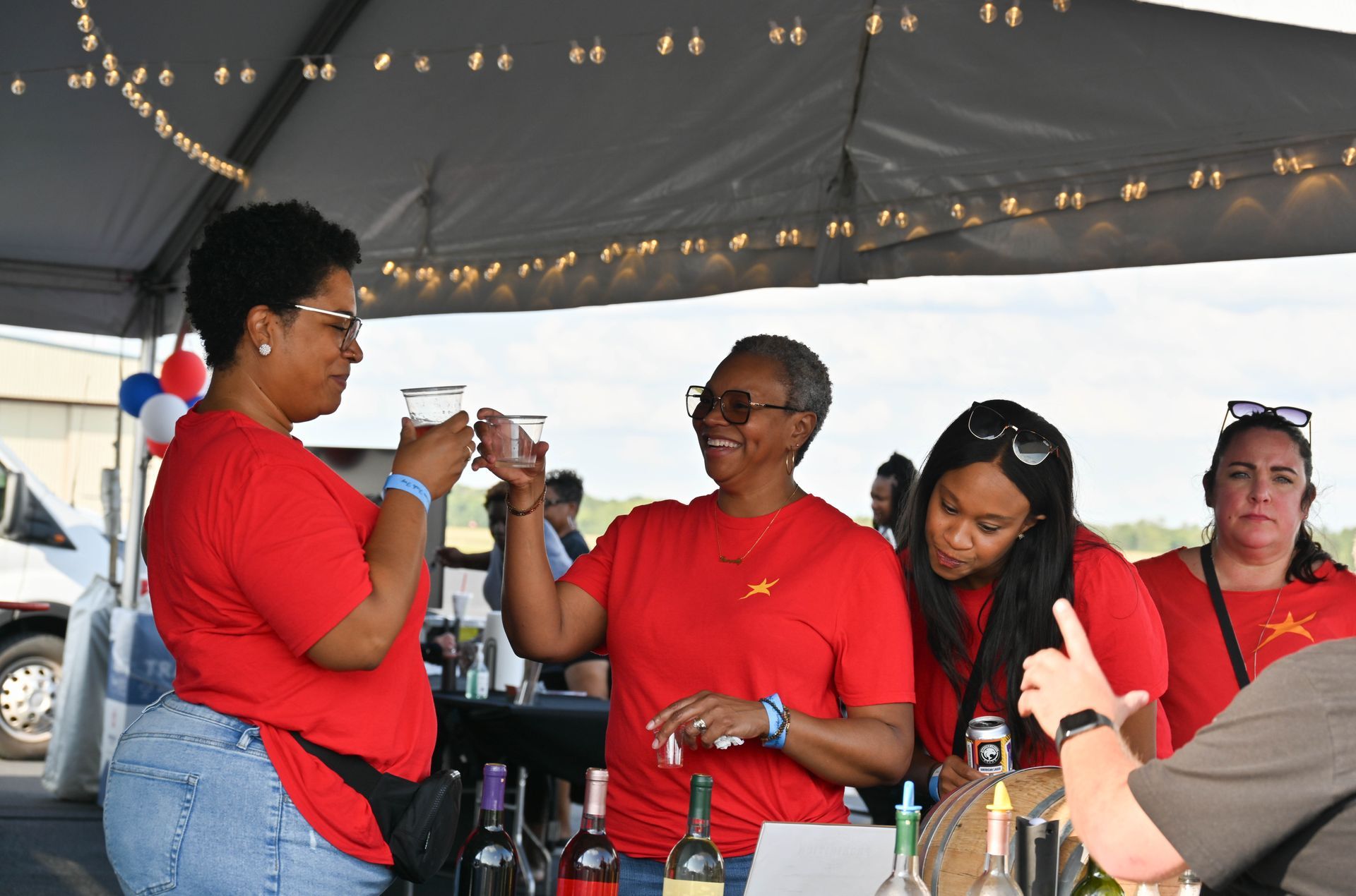 A group of women in red shirts are standing under a tent drinking wine.