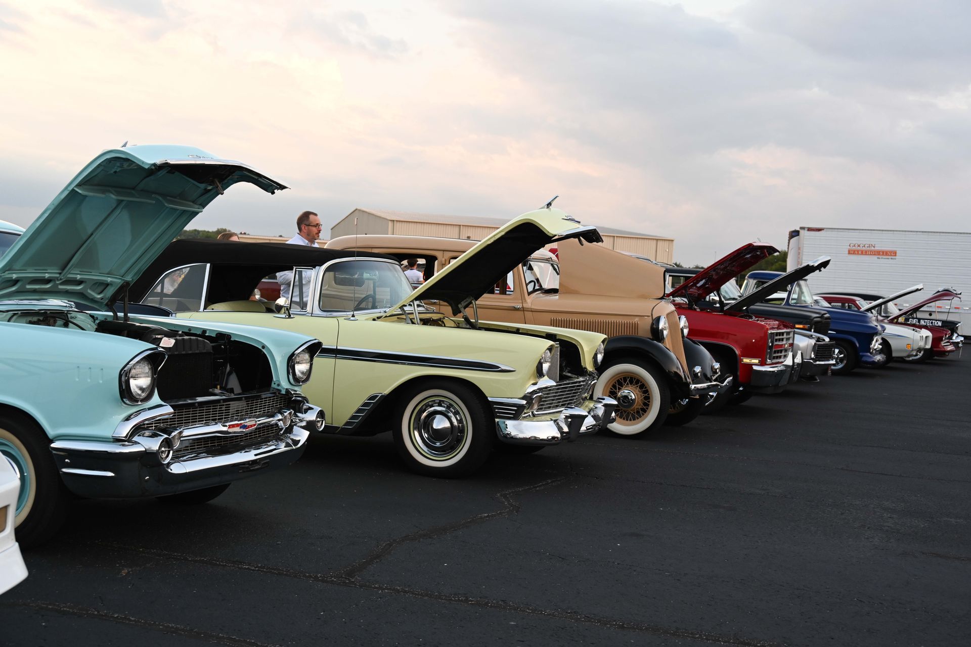 Several old cars are parked in a parking lot with their hoods open