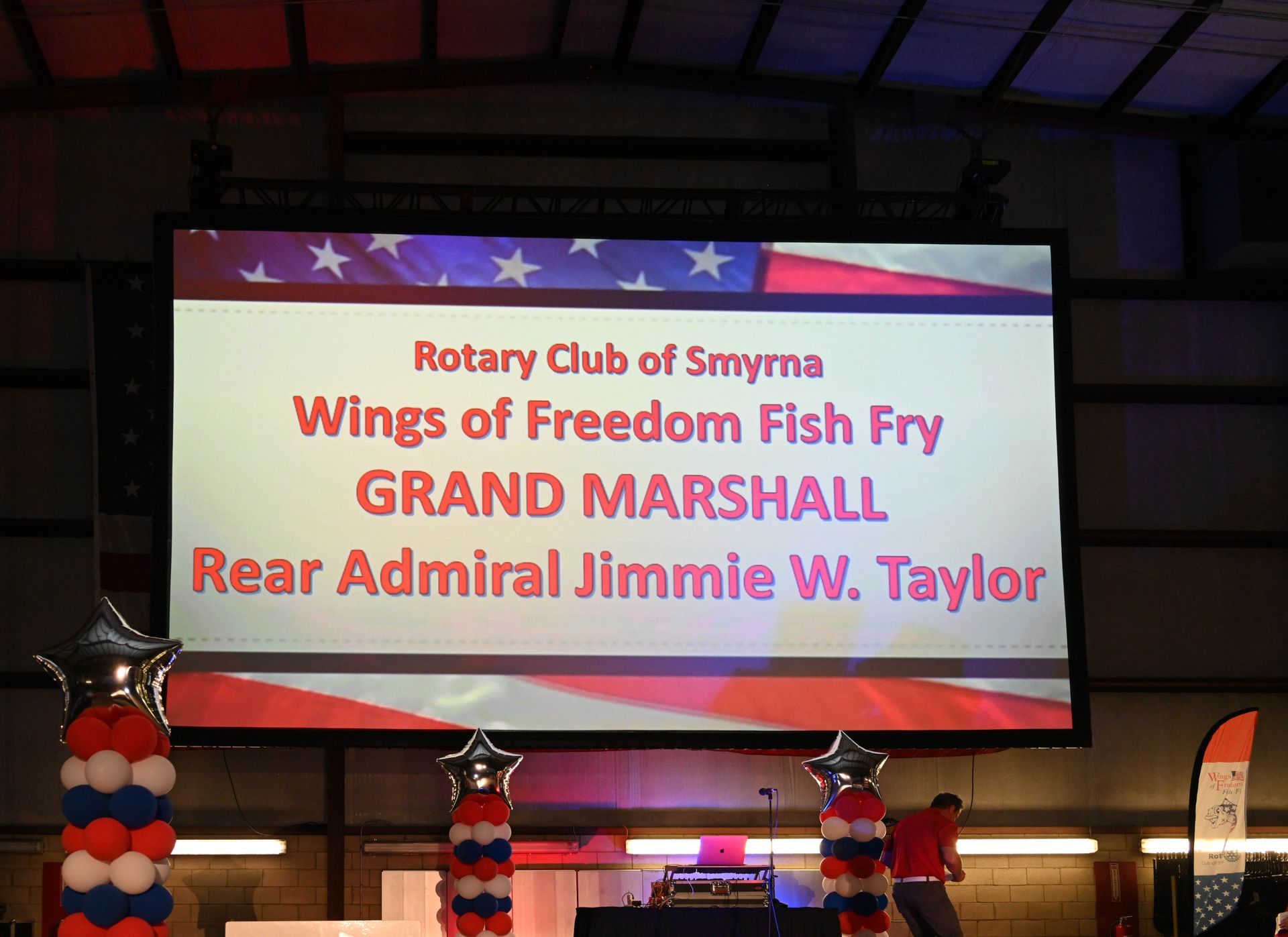 A large screen that says rotary club of smyma wings of freedom fish fry grand marshall rear admiral jimmie w. taylor
