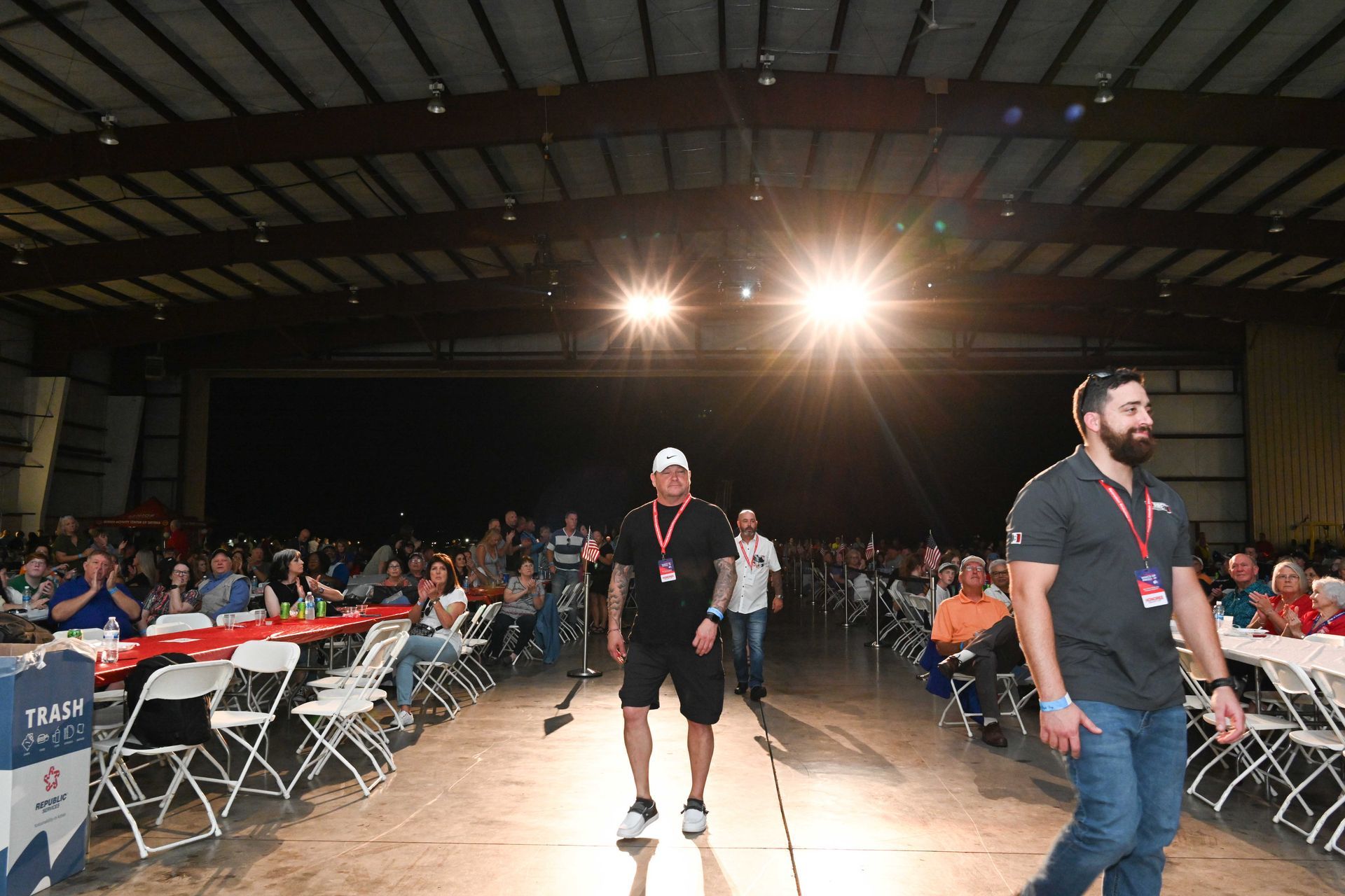 Two men are walking in front of a crowd of people in a large room.