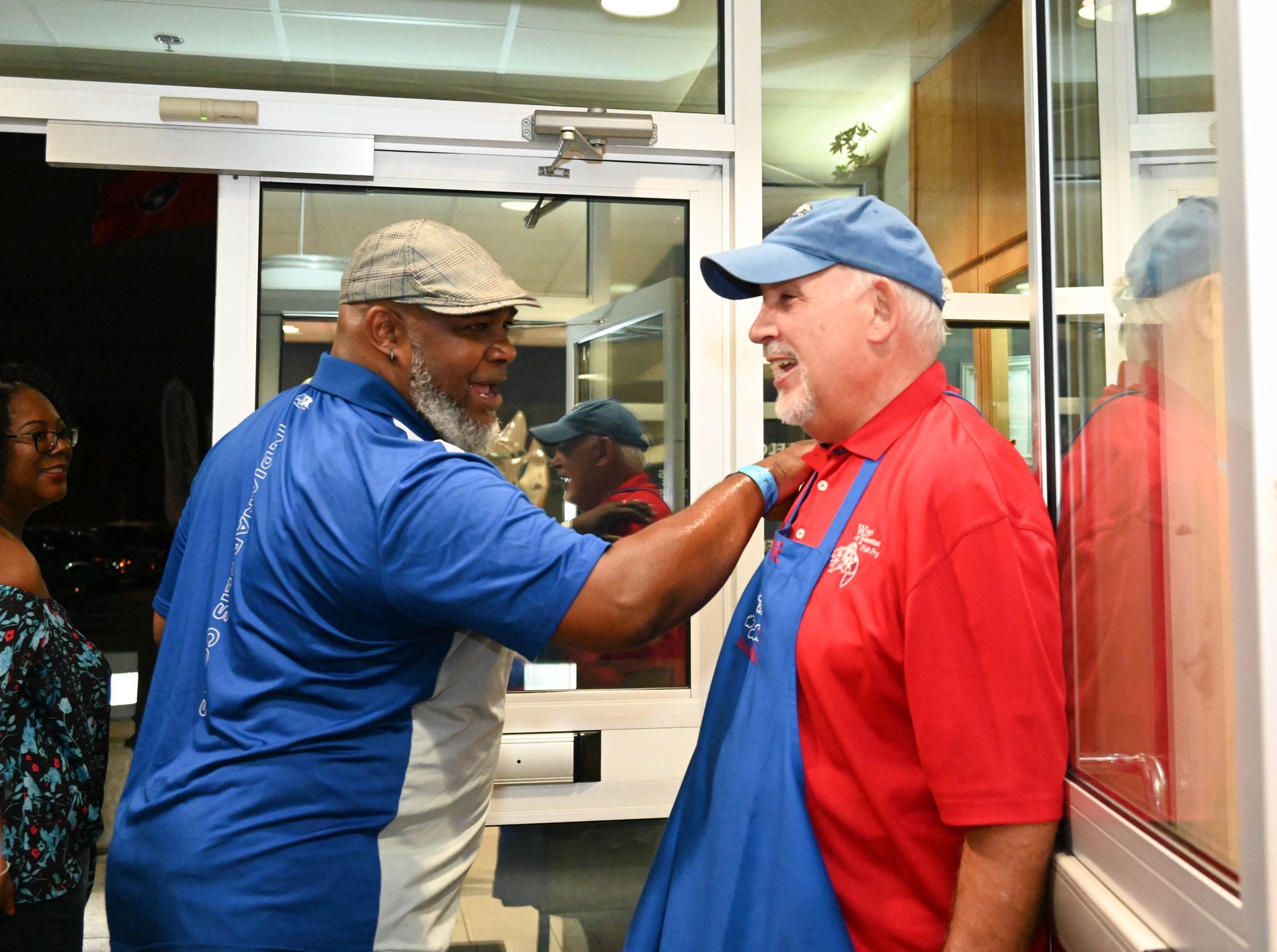 A man in a blue shirt is helping another man in a red shirt with his apron.