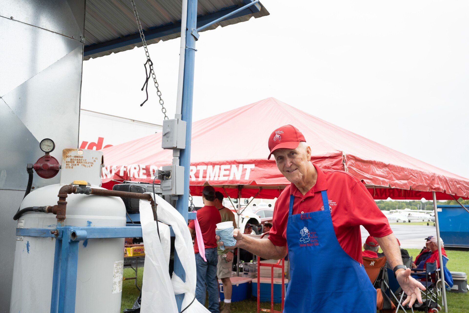 A man in a red shirt and blue apron is standing in front of a red tent.