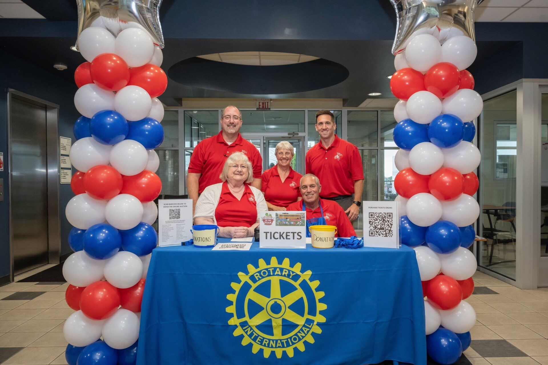 A group of people standing around a table with red , white and blue balloons.