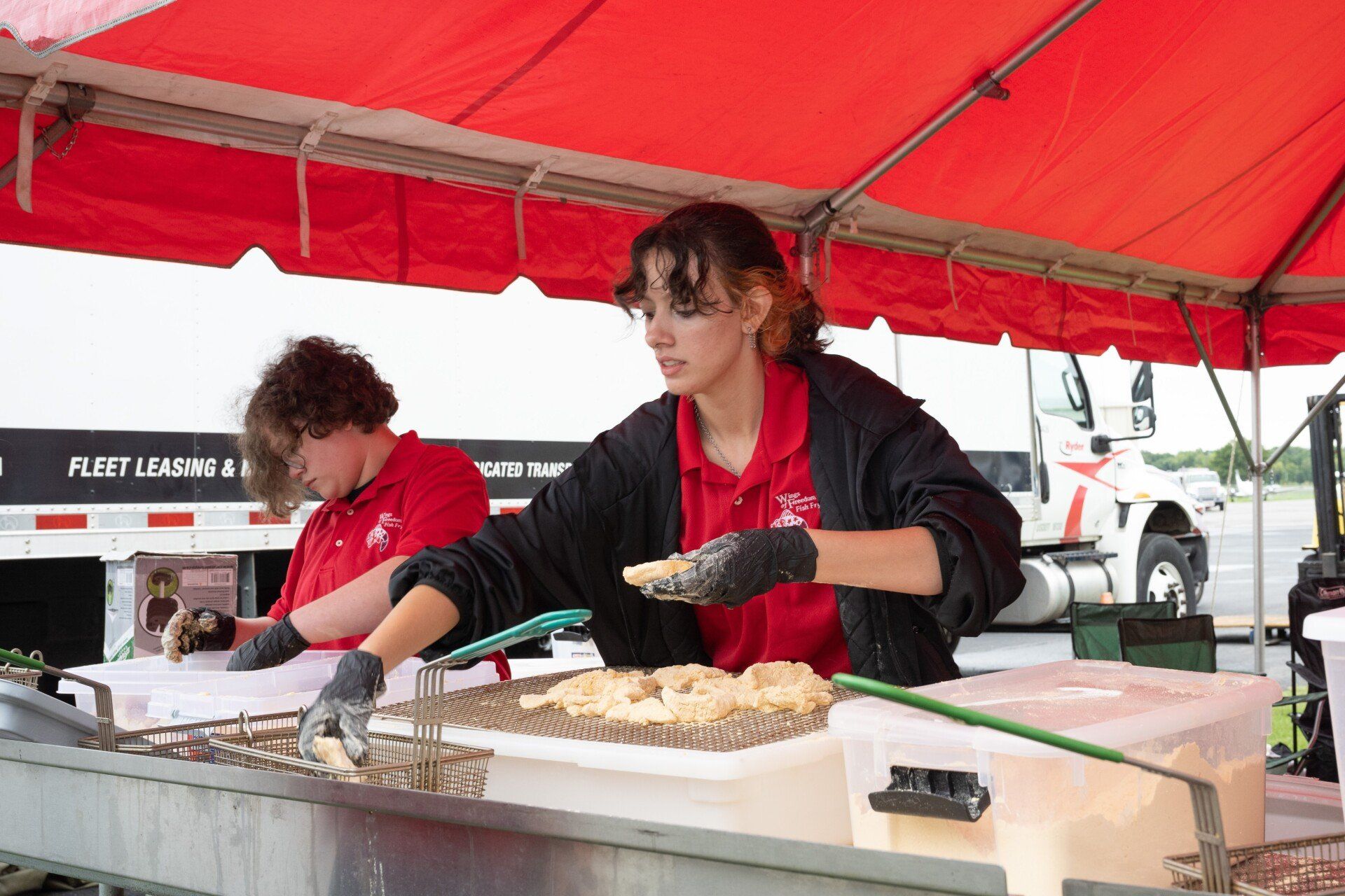 A woman in a red shirt is cooking food under a red tent.