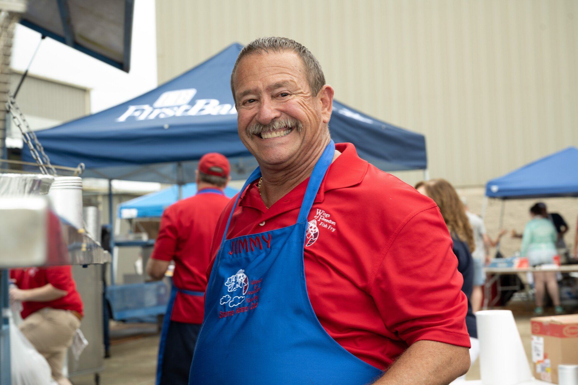 A man in a red shirt and blue apron is smiling at the camera.
