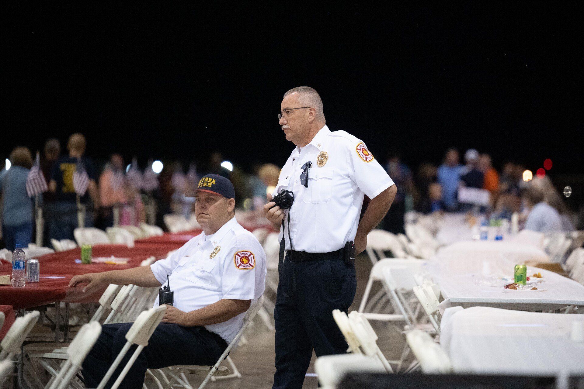 Two men in firefighter uniforms are standing in front of tables and chairs.
