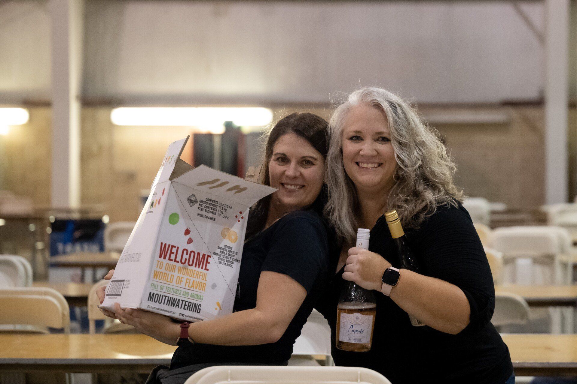 Two women are holding a box and a bottle of wine.