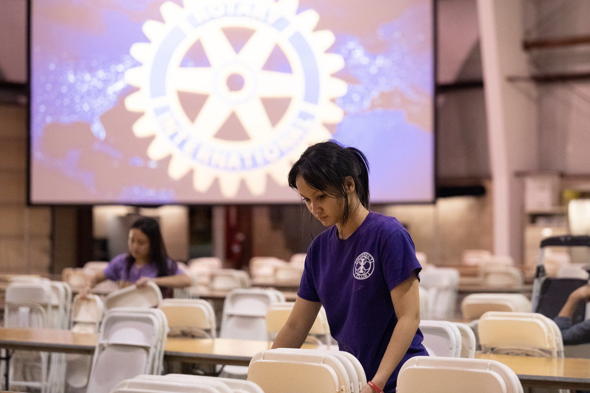 A woman in a purple shirt is arranging chairs in a room with a rotary logo in the background.