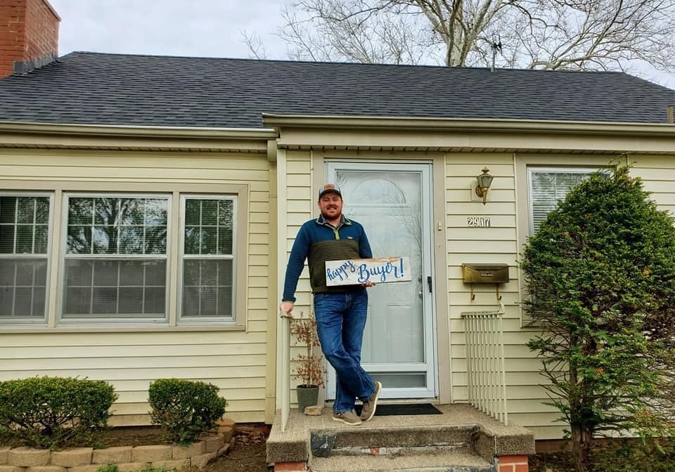 A man is standing in front of a house holding a sign that says work