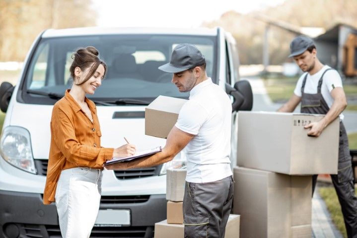 Two movers unloading boxes from a van while a woman checks a clipboard.