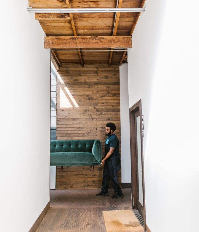 Person carrying a teal sofa through a narrow hallway with wood-paneled walls and a high ceiling