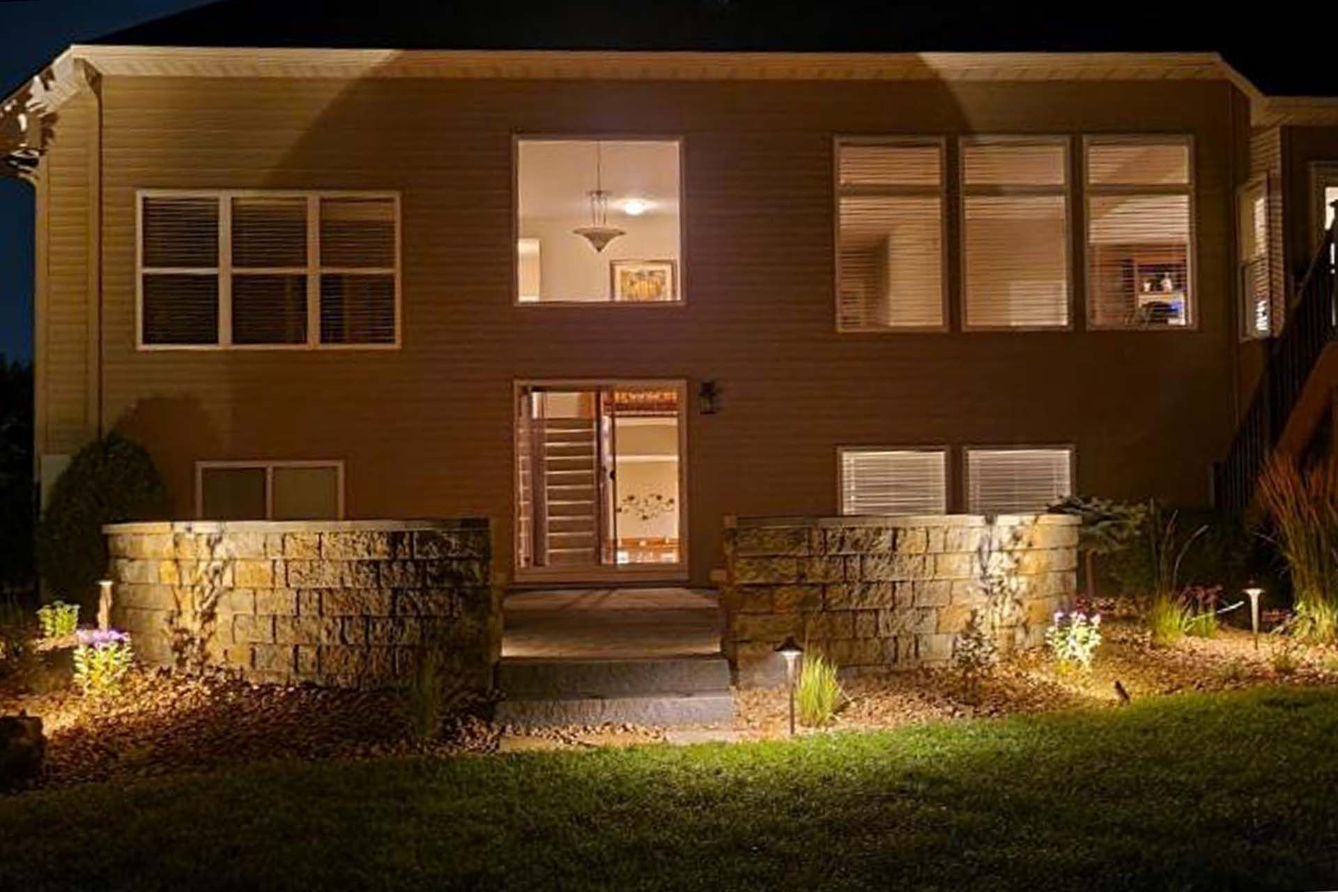 A house with a lot of windows is lit up at night showcasing the retaining wall and lighting.