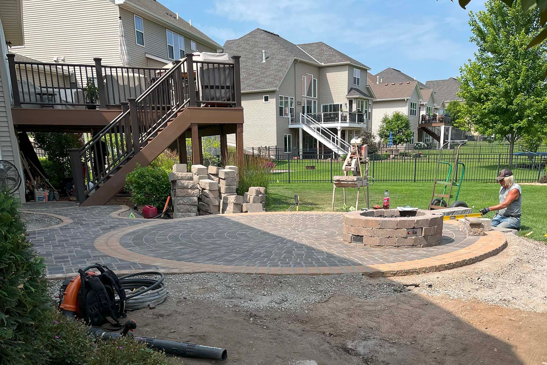 A man is kneeling in front of a fire pit they are building into a patio in a backyard.