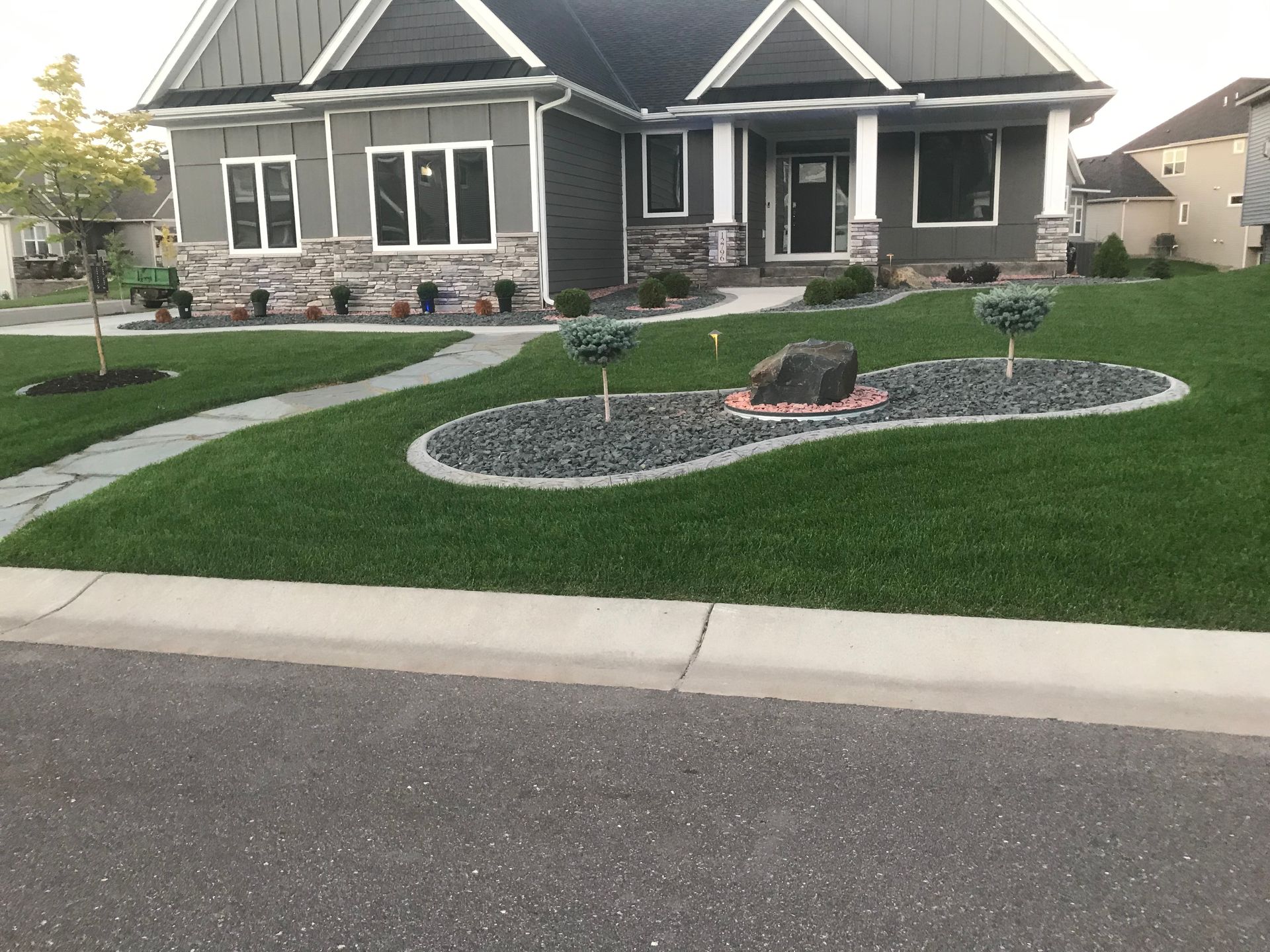A house with a lush green lawn in front of it with a large rock garden with trees. 