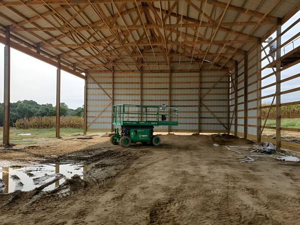 A green scissor lift sits inside an unfinished pole barn with a dirt floor and metal-framed walls.